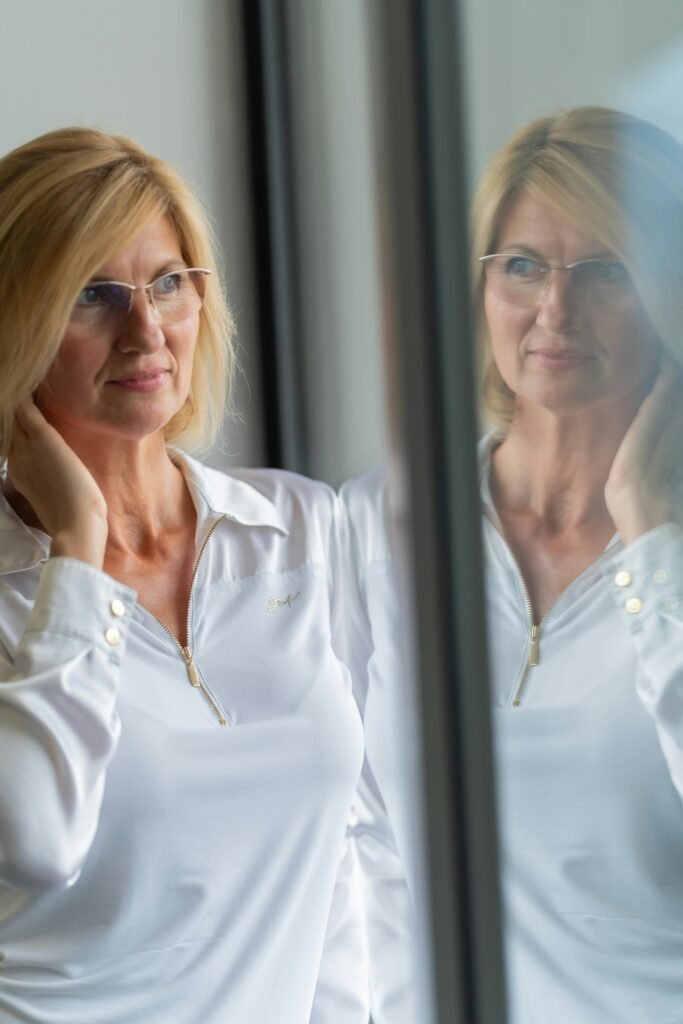Blonde woman in white shirt looking at her reflection in a glass wall indoors.